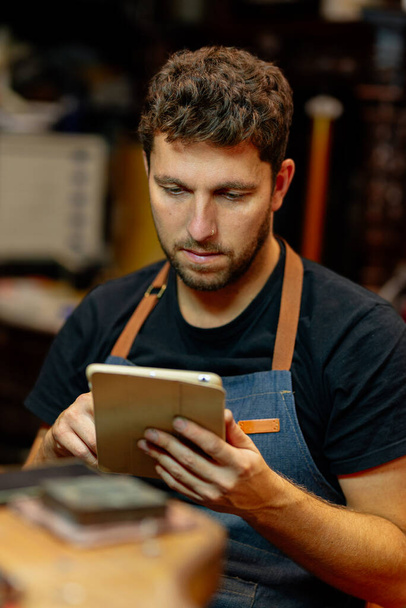 Młody człowiek pracuje w swoim warsztacie, konsultując zamówienia online na tablecie cyfrowym. - Zdjęcie, obraz