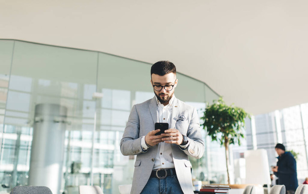 Concentrated male office manager in formal wear messaging on mobile phone while standing in modern building terrace against glass wall on sunny day - 写真・画像