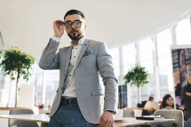 Low angle of serious well dressed male entrepreneur in stylish eyewear standing in modern office in contemporary business centre at  daytime - 写真・画像