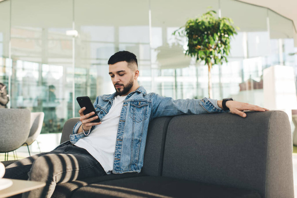 Young handsome male in denim jacket and white T shirt surfing social media on cellphone while sitting on sofa in modern shopping mall - Photo, Image