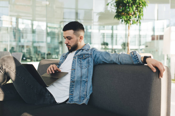 Side view of serious bearded enterpreneur in stylish clothes sitting on comfortable sofa in modern workspace and using laptop while working on project - Photo, image
