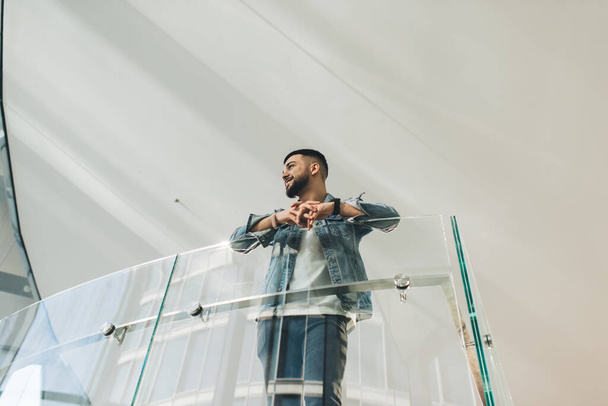 Снизу мужчина в джинсовой куртке смотрит в сторону и опирается на перила, стоя в коридоре креативного здания - Фото, изображение