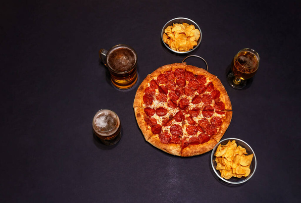 Lahodná feferonková pizza, sklenice piva, hrnek piva a bramborové lupínky na tmavém pozadí. Horní pohled, kopírovací prostor. - Fotografie, Obrázek
