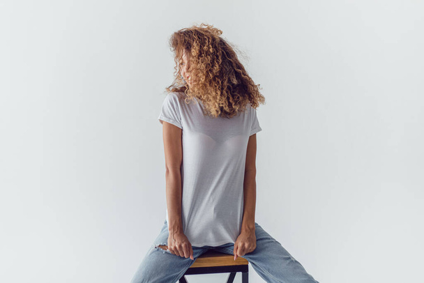 Smiling woman with lush curly hair in white blank t-shirt sits on a white background. Mock-up. - Φωτογραφία, εικόνα