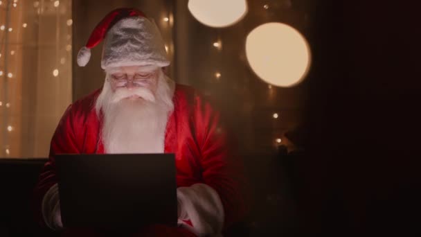 De kerstman werkt 's nachts met een laptop in het licht van kerstverlichting - Video