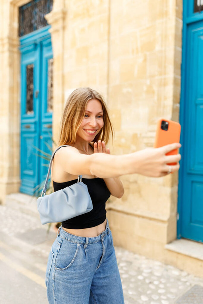 Młoda piękna dziewczyna w dżinsach i torebce robi selfie na tle cypryjskiego starego miasta - Zdjęcie, obraz