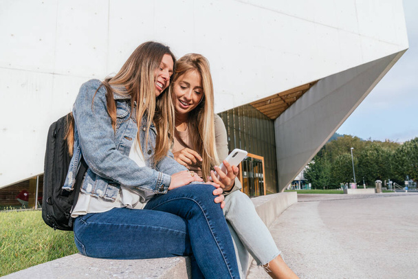 Side view of two female college classmates sitting near of university using phone and having fun. - Photo, image