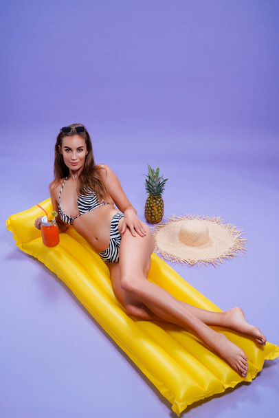 happy woman wears in bikini lyes on inflatable matress while holds cocktail on purple background  - Foto, imagen