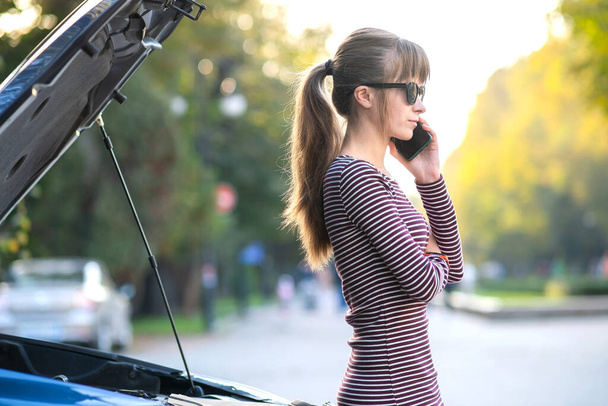 Jonge overstuur vrouw bestuurder praten op mobiele telefoon in de buurt van een kapotte auto met open motorkap wachten op hulp hebben met haar voertuig in een stad straat - Foto, afbeelding