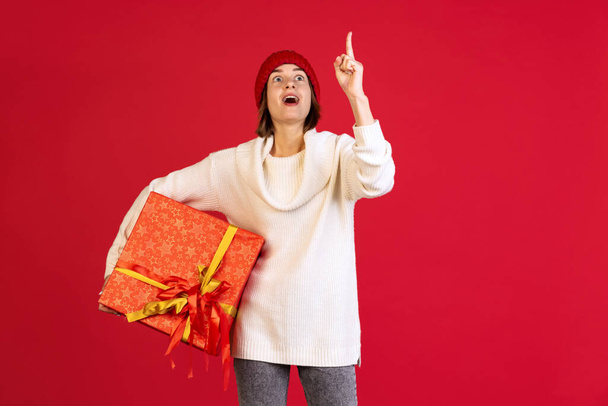 Cropped portrait of excited young woman with big present box pointing with finger up isolated over red background. Gift ideas - Foto, Imagem