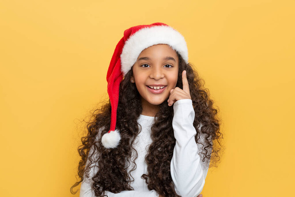 Waist up portrait of a dark skinned young girl with long curly hair in a Santa Claus hat on a yellow background - Fotó, kép