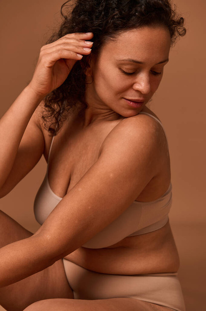 Portrait of a dark-haired curly woman with skin flaws, cellulite and stretch marks posing in beige underwear, looking down against colored background with copy space. Body positive concept - Fotografie, Obrázek