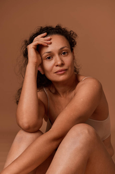 Portrait studio d'une femme d'âge moyen aux cheveux bruns en sous-vêtements regardant avec confiance la caméra, posant sur un fond de couleur beige avec un espace de copie pour la publicité. Positivité corporelle - Photo, image