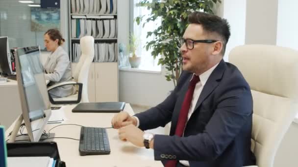 Porträt eines glücklichen männlichen Managers in Anzug und Brille, der am Schreibtisch im Büro sitzt und die Arbeit am Computer beendet, sich dann der Kamera zuwendet und in seinem Stuhl lächelt - Filmmaterial, Video