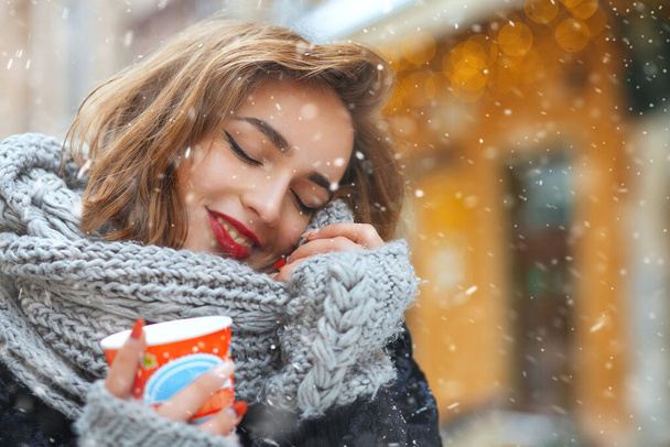 Álmodozó fiatal nő egy csésze kávéval sétál a díszített vitrinek közelében havas időben. Üres tér - Fotó, kép