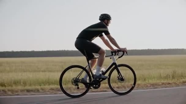 Cyklista jezdí na silničním kole při západu slunce na dálnici u pole, nosí helmu - Záběry, video