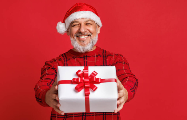 赤いサンタの帽子を持つ陽気な驚きの先輩はクリスマスの贈り物を保持し、色の赤い背景を笑って - 写真・画像