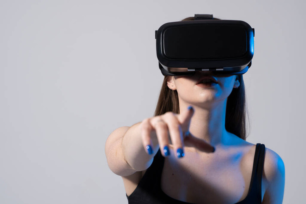 Brunette woman in a black t-shirt and VR headset looking up and trying to touch objects in virtual reality. VR is a technology that simulates a physical presence and to interact with environment. - Foto, Imagem