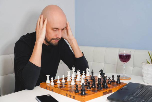 Homme chauve réfléchi portant la barbe jouant aux échecs à bord près d'un ordinateur portable et du verre à vin. Stratégie et concept de concurrence. Vue latérale - Photo, image