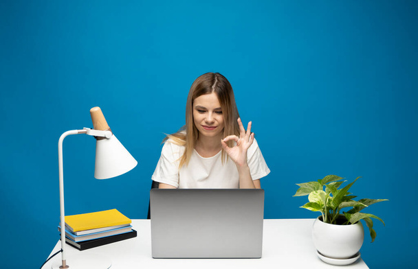 Its okay. Overjoyed woman in a white shirt and glasses showing ok sign, sitting with laptop over blue background. - Фото, изображение