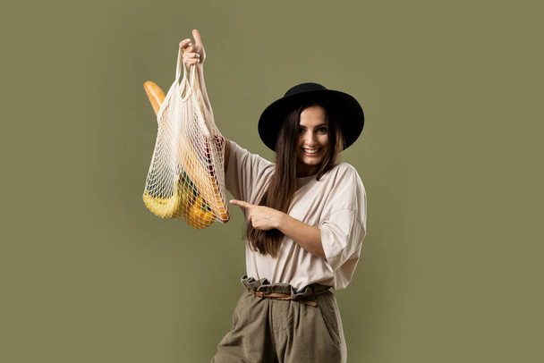 Mesh eco bag with groceries, vegetables and fruits, in the hands of a brunette girl in a beige t-shirt and black hat. Eco friendly concept. Woman holding string shopping bag. - 写真・画像