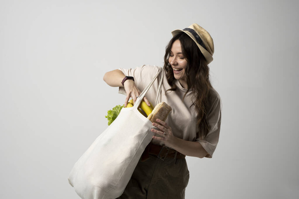 Smiling woman in a t-shirt and a hat holding reusable cotton shopping bag with vegetables, bread and greens. Concept of no plastic, zero waste, plastic free, sustainable lifestyle. - Foto, imagen