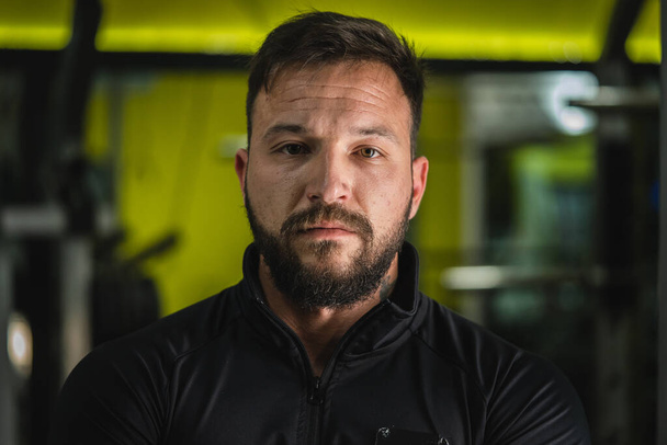 Front view close up portrait of young adult caucasian man with dark hair and beard in black gym suit looking to the camera in dark - Confident modern male dramatic portrait with copy space - Photo, Image