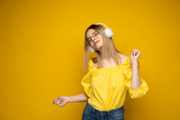 Happy girl in a glasses and yellow shirt dancing and listening to the music isolated on a yellow background. - Fotografie, Obrázek