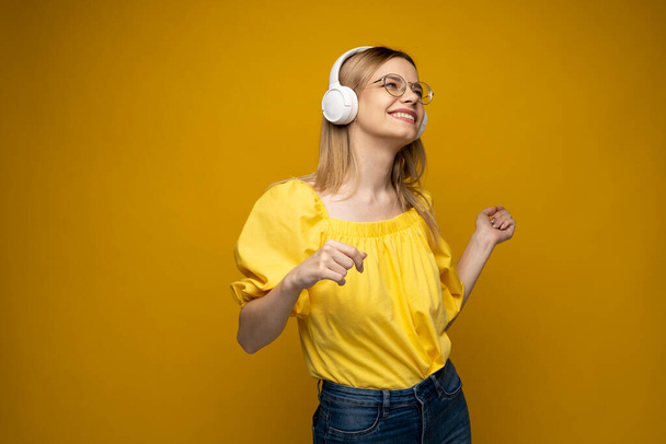 Wesoła młoda ładna dziewczyna uśmiecha się słuchając muzyki w słuchawkach i tańcząc na żółtym tle. - Zdjęcie, obraz