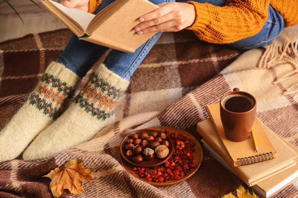 Mujer en calcetines calientes con libro, taza de café, comida y hojas de otoño sobre fondo de tela a cuadros - Foto, Imagen