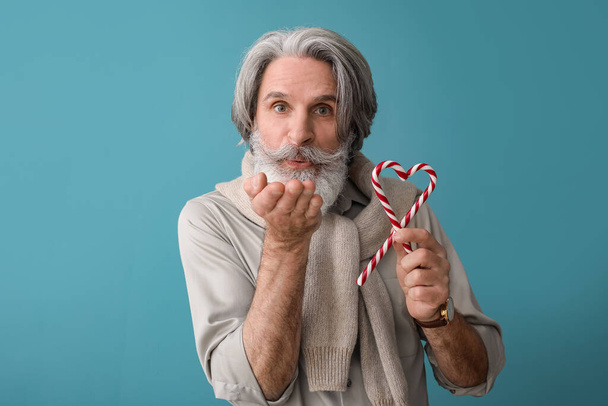 Senior man with sweet candy canes blowing kiss on color background - Photo, Image