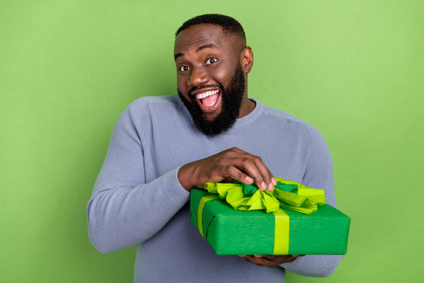 Photo of funny african guy dressed grey sweater smiling opening gift package isolated green color background - Photo, image
