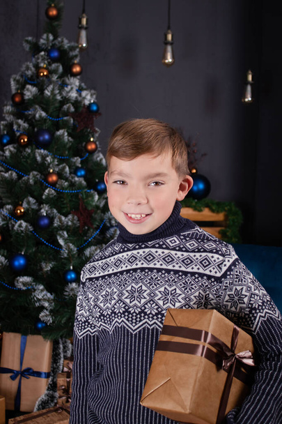 Boldog, csinos szőke fiú kötött pulóverben, karácsonyi ajándékkal egy karácsonyfa mellett. Újévi ünneplés. - Fotó, kép