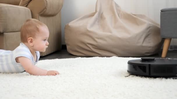 小さな赤ちゃんの男の子とロボット掃除機が近づいてきて、リビングルームのカーペットの上に近づく。現代生活における衛生、家庭用品、ロボットの概念. - 映像、動画
