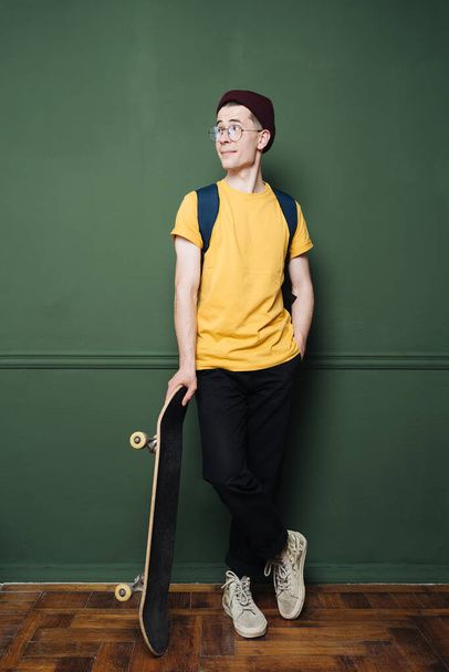 Caucasian man in yellow t shirt posing with skate over dark green background wall - Foto, afbeelding