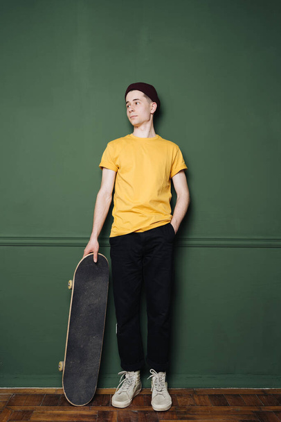 Homem caucasiano em t-shirt amarela posando com skate sobre parede de fundo verde escuro - Foto, Imagem