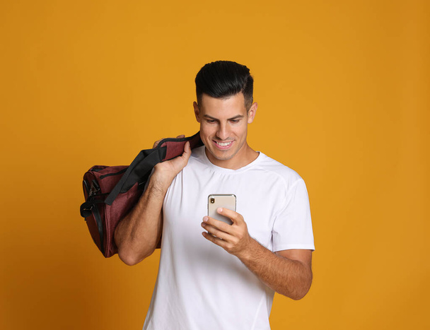 Handsome man with sports bag and smartphone on yellow background - Fotoğraf, Görsel