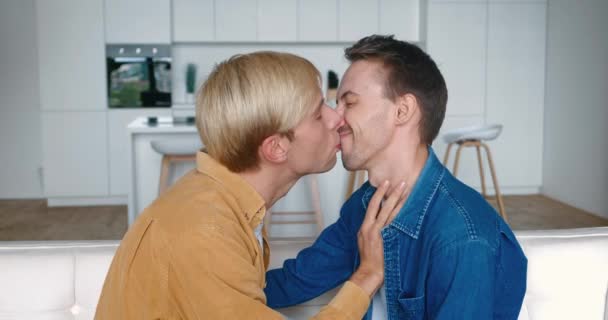Portret dwóch szczęśliwych mężczyzn gejów para w miłości całowanie w zrobić delikatny tyłek podczas siedzenia na kanapie w domu. Stosunki LGBT - Materiał filmowy, wideo