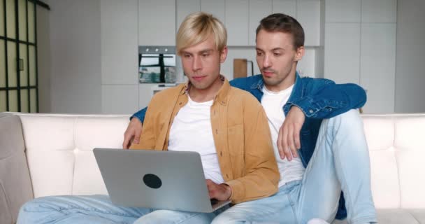 wstrząśnięty mężczyzna gej para patrząc w laptop ekran i zdenerwowany z złe wiadomości podczas siedzi razem na kanapie w domu - Materiał filmowy, wideo