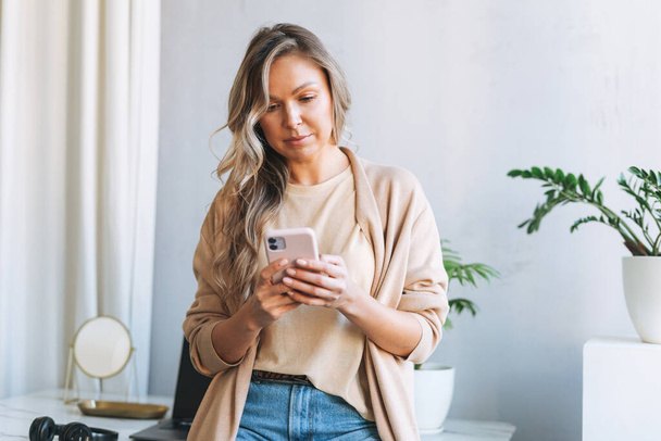 Young smiling blonde woman with long hair in stylish cardigan working at laptop in bright modern office, woman send message by smartphone - Photo, image