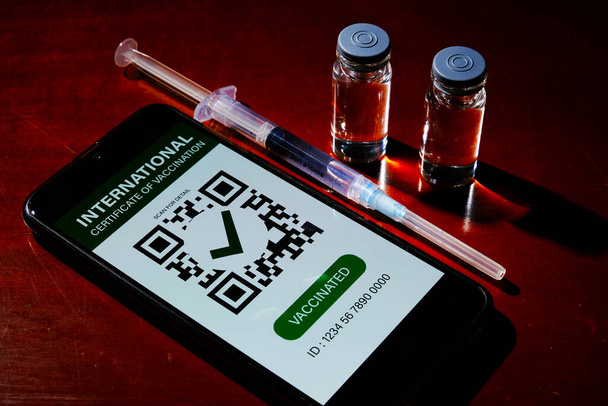 Uma foto no modo escuro do smartphone com falso Certificado Internacional de Vacinação nele, duas doses de vacina e insights de seringa para injeção na mesa. Dose após completar 2 doses - Foto, Imagem
