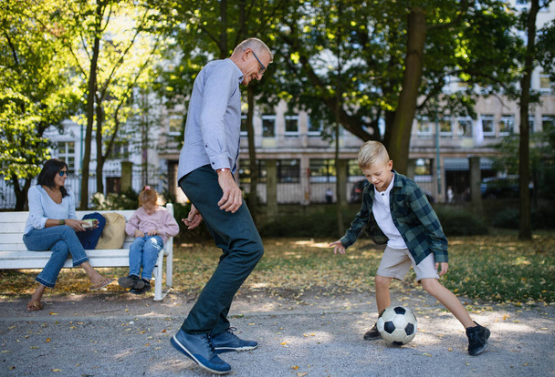 Little boy playing football with granddad, his sister and grandmom sitting on bench outdoors in park - Fotoğraf, Görsel