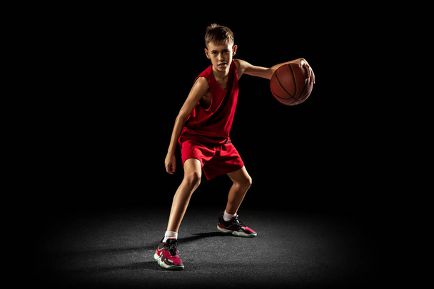 Full-length portrait of teen boy, professional backetball player in motion, training isolated over black background. Dribbling action - Photo, Image