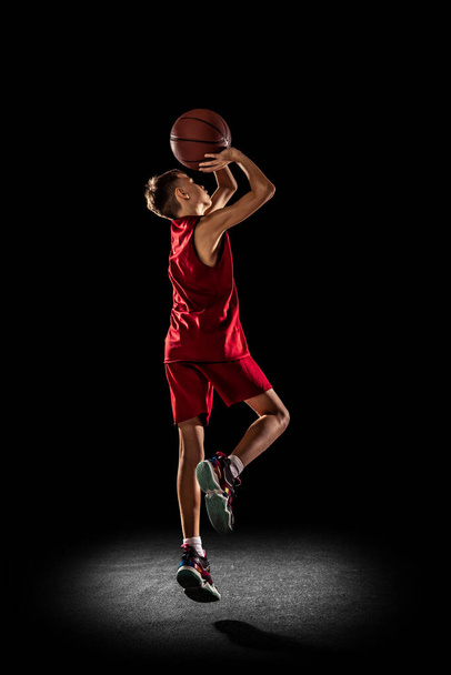 Ganzkörperporträt eines sportlichen Teenagers, Basketballspielers in roter Uniform, der Ball isoliert über schwarzem Hintergrund in den Korb wirft - Foto, Bild