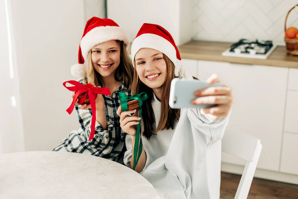 Portret van twee schattige zussen in een kerstman Kerstmuts in de keuken, selfies nemen met een smartphone, terwijl de voorbereiding van geschenken op kerstavond en oudejaarsavond. - Foto, afbeelding