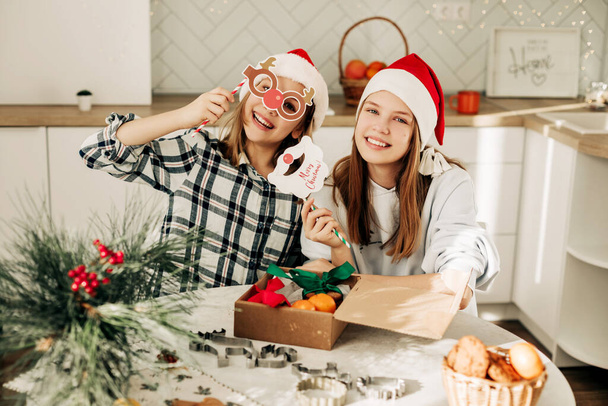 Δύο κορίτσια με χριστουγεννιάτικα καπέλα πακετάρουν χριστουγεννιάτικα δώρα, οι αδερφές διασκεδάζουν με Χριστουγεννιάτικες μάσκες.. - Φωτογραφία, εικόνα