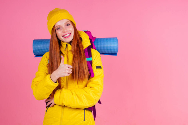 Female hiker with a backpack looking at the camera and smiling isolated on pink studio background.she wearing yellow windbreaker waterproof jacket and knitted hat - Fotó, kép