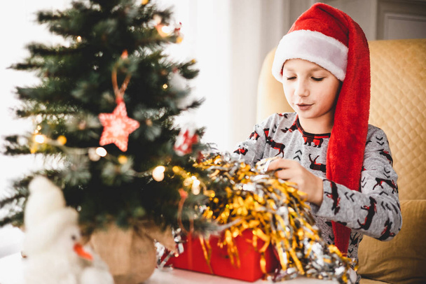 Święta i Nowy Rok. Szczęśliwy chłopiec w piżamie i czerwony kapelusz Mikołaja otwierający pudełko w domu wczesnym rankiem. Duch wakacji zimowych - Zdjęcie, obraz