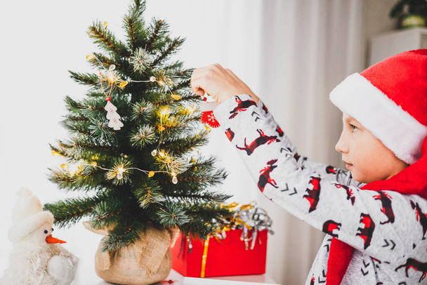 Vánoce a novoroční prázdniny. Malý chlapec zdobí malý vánoční stromeček během zimních prázdnin. Teenager v pyžamu a červené Santa klobouk závěsné dekorativní dřevěné hračky na jedlových větvích. - Fotografie, Obrázek