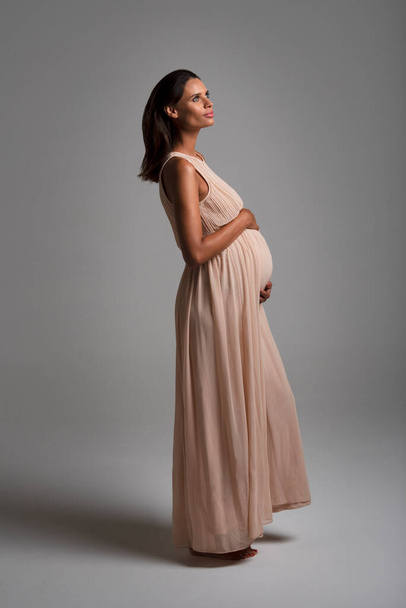 Mladá krásná těhotná žena stojící na podlaze na sobě dlouhé růžové šaty vertikální pohled šedé pozadí - Fotografie, Obrázek